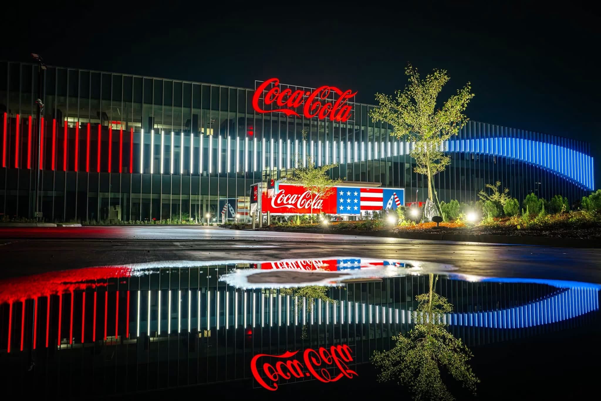 Coca-Cola Birmingham plant illuminated at night with red, white, and blue LED accent lighting reflected in a foreground puddle