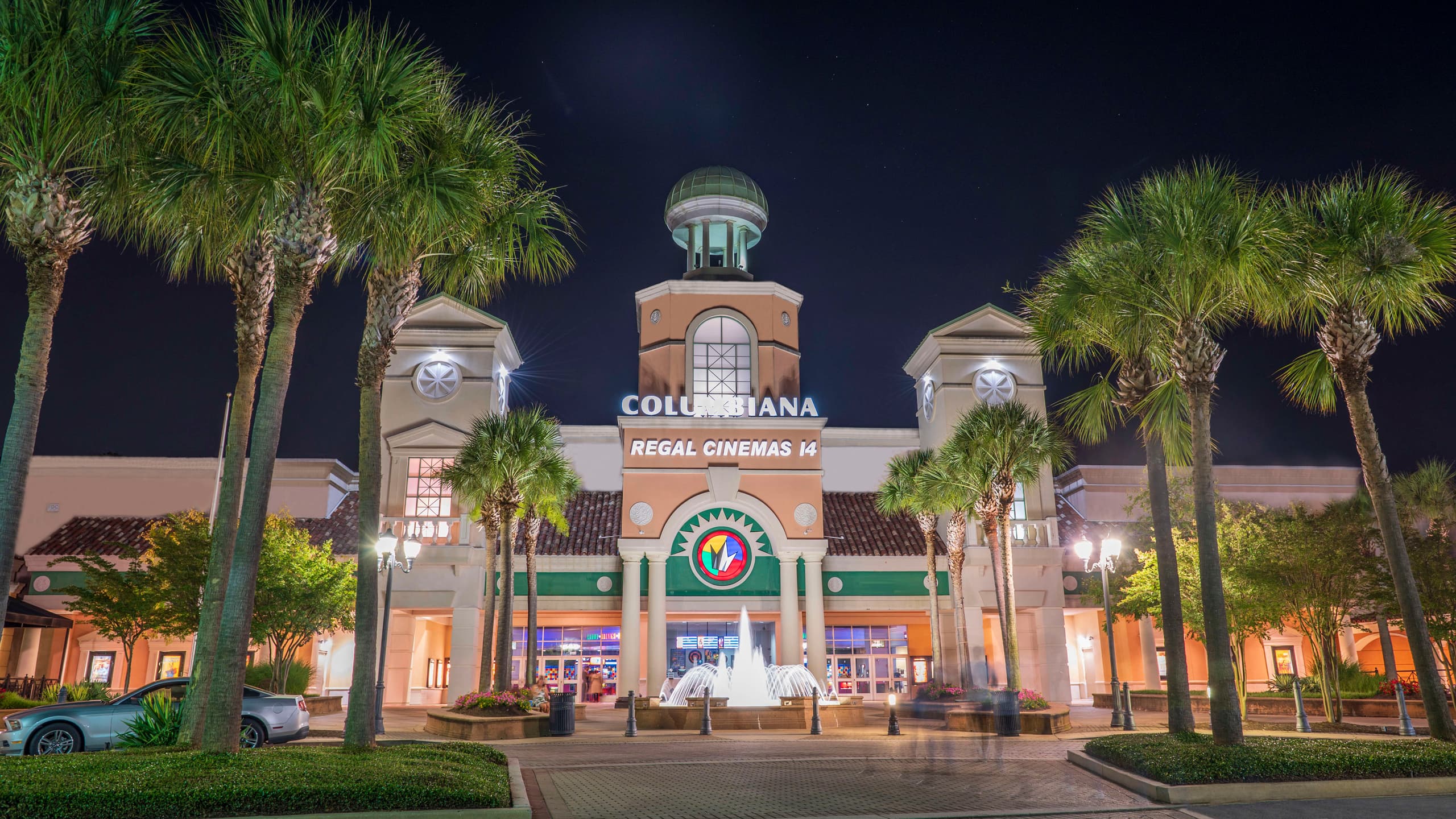 Columbiana Station shopping center at night, its entrance tower, palms, and storefronts evenly lit