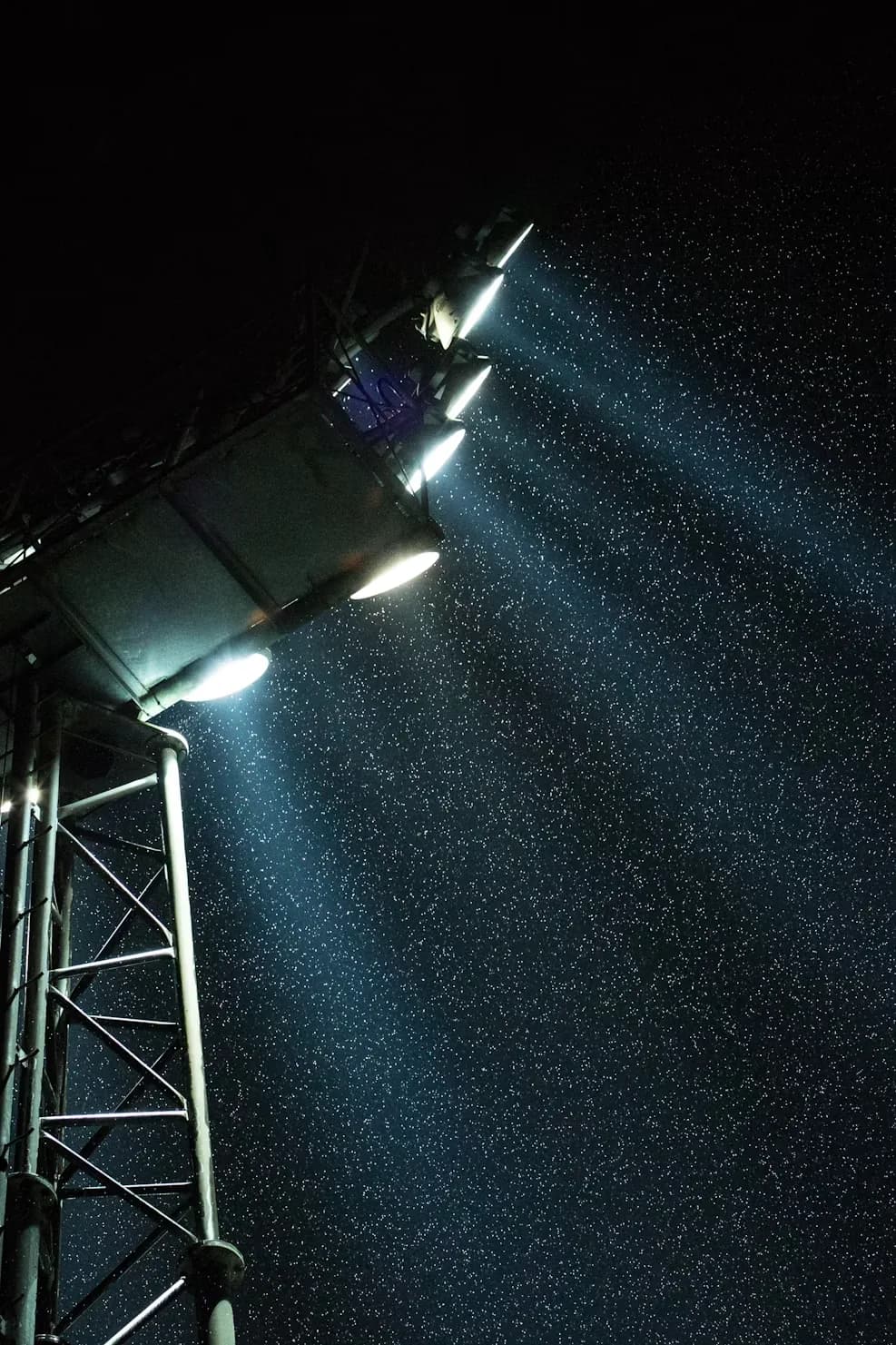 A floodlight head seen close and from below at night, its beam cutting through falling rain.