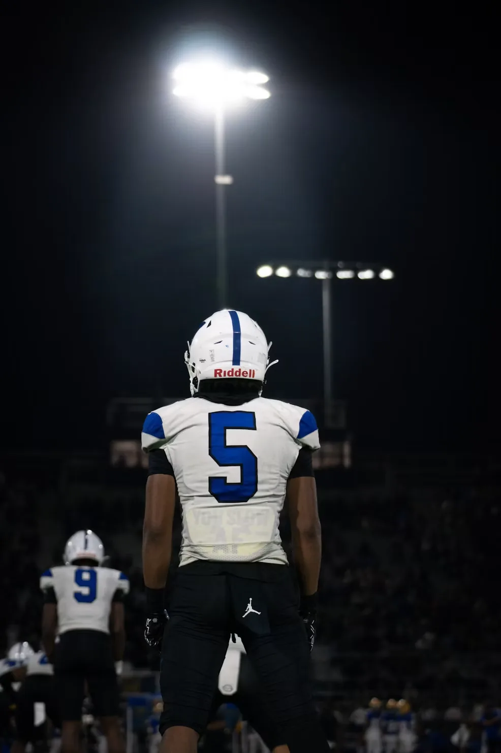 A football player seen from behind under floodlights at night, the lit field ahead of him.