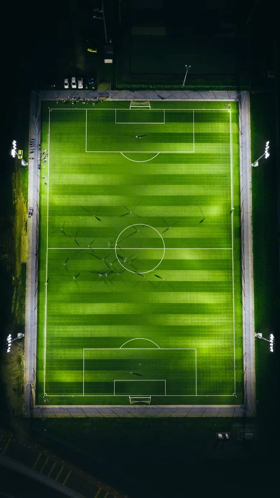 A floodlit soccer pitch from directly overhead at night, masts at the corners, mowing stripes visible.